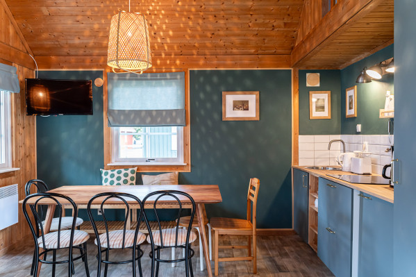 Gemütlicher Essbereich mit Holztisch, modernen Stühlen und Fernseher, angrenzend an eine blau gestaltete Küchenzeile mit Holzdetails.